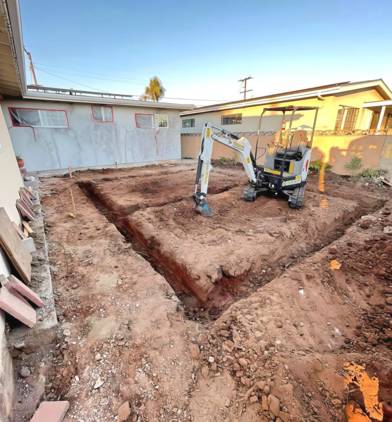 A mini excavator sits in a residential backyard with freshly dug trenches forming an L-shape pattern in reddish-brown soil. The construction site is surrounded by houses with one structure covered in protective sheeting, suggesting renovation work in progress. The scene is captured during golden hour under clear blue skies.