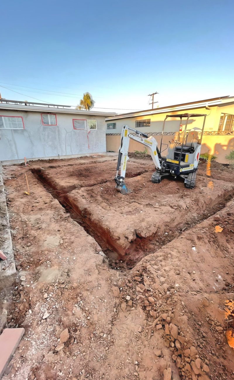 A mini excavator sits in a residential backyard with freshly dug trenches forming an L-shape pattern in reddish-brown soil. The construction site is surrounded by houses with one structure covered in protective sheeting, suggesting renovation work in progress. The scene is captured during golden hour under clear blue skies.