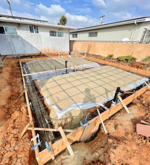 A concrete foundation under construction in a residential backyard, showing two rectangular slabs with rebar reinforcement grids and wooden formwork frames. The freshly poured concrete is surrounded by reddish-brown excavated soil, with white residential buildings visible in the background under a partly cloudy sky. Blue painter's tape marks various points on the wooden forms, and construction materials are scattered around the work site.