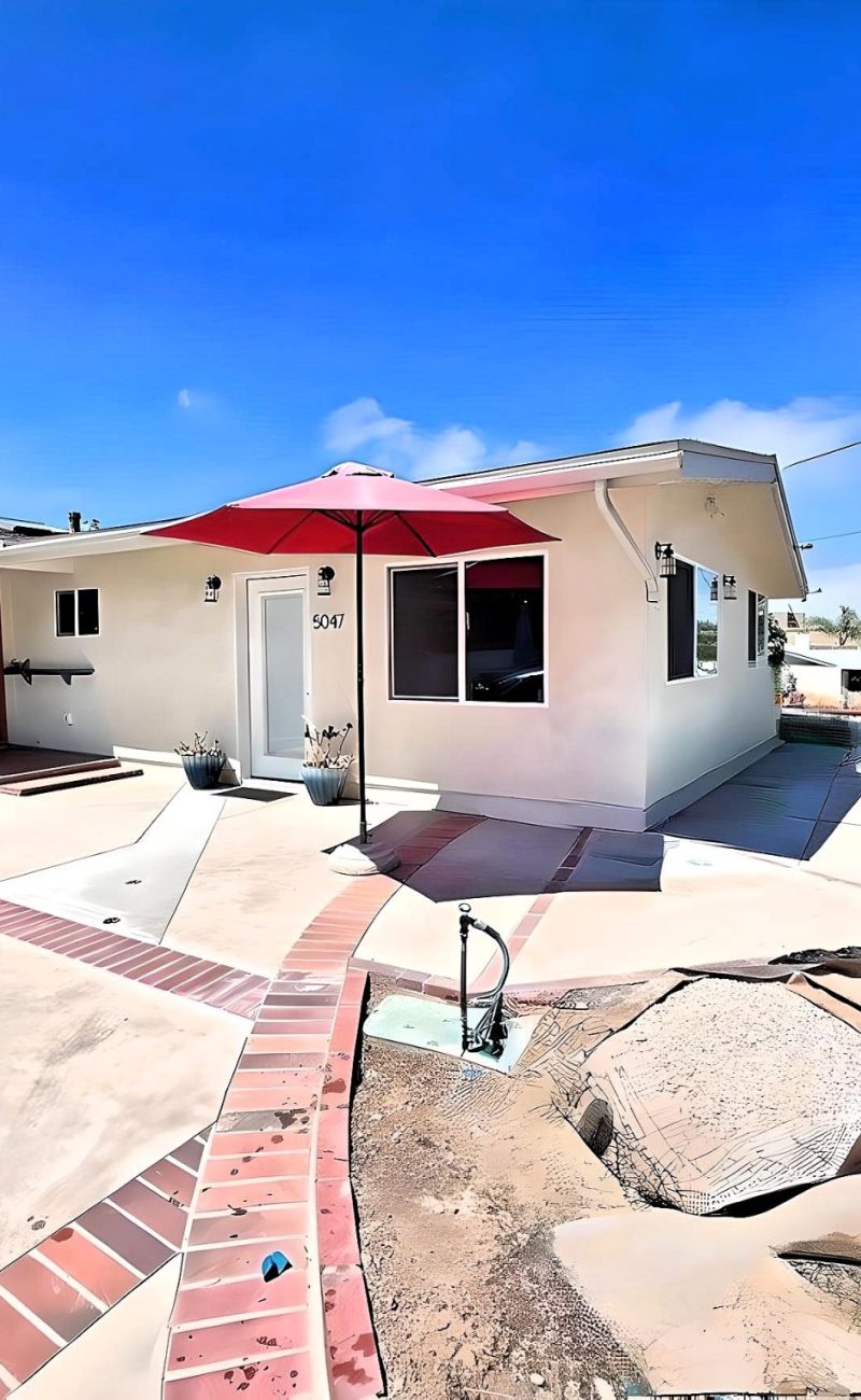 A single-story ranch-style home with beige stucco exterior featuring a front patio area with red brick accents and borders. The entrance has a red patio umbrella, and the property includes a large concrete driveway or courtyard space with decorative brick trim patterns. The home is situated on a corner lot with views of the surrounding residential neighborhood under a bright blue sky.