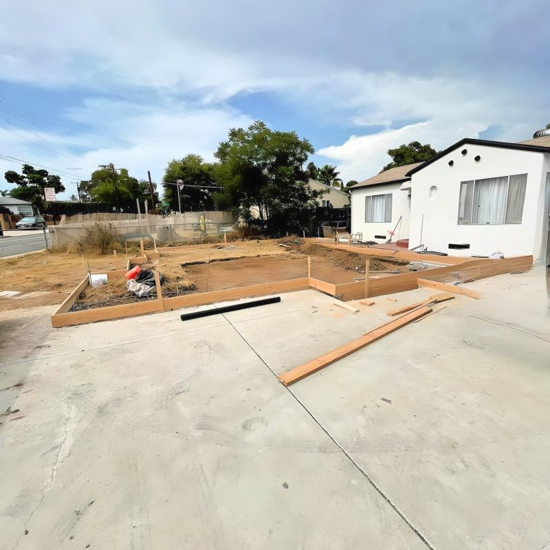 A residential construction site showing wooden frame forms laid out on bare dirt and concrete foundation in preparation for building. A white single-story house with Spanish-style architecture is visible on the right side of the property. The scene includes a cloudy sky, surrounding trees, and utility poles in a suburban neighborhood setting.