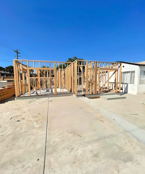 A residential construction site showing wooden framing for a home addition or new structure on a concrete slab foundation. The bare wood studs and wall frames are visible against a clear blue sky, with an existing white building and utility pole in the background. The concrete pad shows typical construction staging with visible seams and weathering.