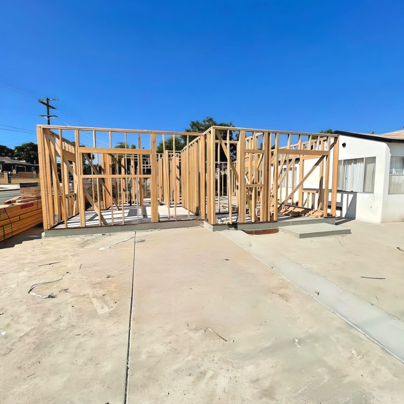 A residential construction site showing wooden wall framing in progress on a concrete foundation. The skeletal structure reveals the layout of future rooms, with vertical studs and headers forming doorways and wall sections. An existing white stucco building is visible on the right side, and the scene is set under a clear blue sky.