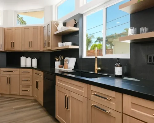 A modern kitchen featuring natural wood cabinets with black hardware paired with sleek black countertops and a matching black backsplash. Large white-framed windows allow natural light to flood the space and provide views of palm trees outside. The design includes open wooden shelving on the wall and wood-toned flooring, creating a warm contemporary aesthetic with a black and natural wood color scheme.