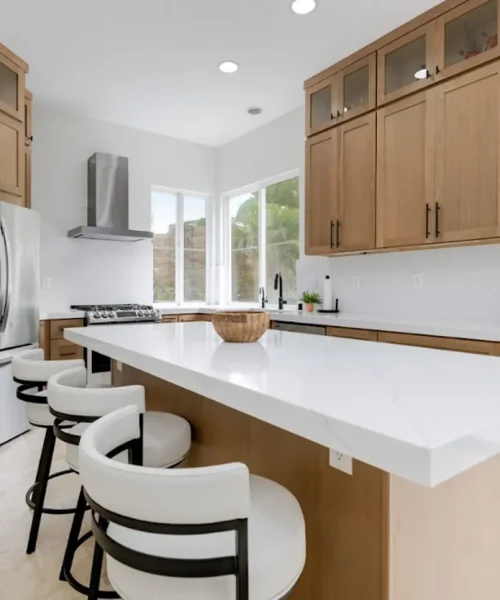 A modern kitchen featuring light wood cabinetry with black hardware, white countertops, and a large island with white upholstered bar stools. The space includes stainless steel appliances, a stainless range hood, and bright natural light from multiple windows overlooking greenery. The design showcases a clean, contemporary aesthetic with a warm neutral color palette combining natural wood tones and white surfaces.