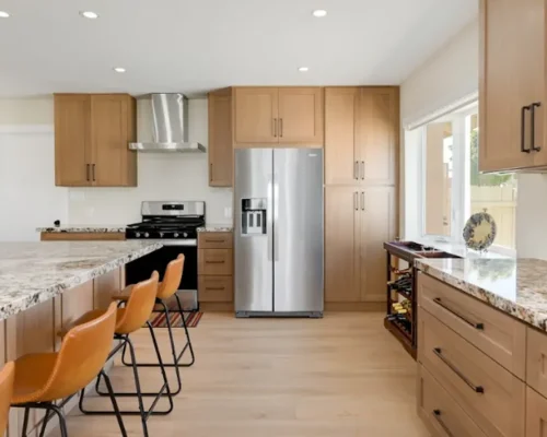 A modern kitchen featuring light wood cabinetry with dark hardware, granite countertops, and stainless steel appliances including a French door refrigerator and gas range with overhead hood. Three orange-brown bar stools with black metal frames are positioned at a kitchen island. The space has light wood flooring, white walls, recessed lighting, and a window providing natural light.