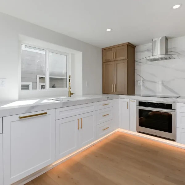 A modern kitchen featuring white shaker-style cabinets with gold brass hardware and light wood accents. The space includes a marble backsplash, built-in stainless steel appliances, floating wooden shelves, and distinctive orange LED lighting underneath the lower cabinets that illuminates the light hardwood flooring.