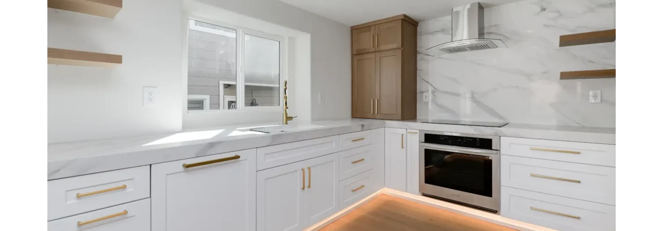 A modern kitchen featuring white shaker-style cabinets with gold brass hardware and light wood accents. The space includes a marble backsplash, built-in stainless steel appliances, floating wooden shelves, and distinctive orange LED lighting underneath the lower cabinets that illuminates the light hardwood flooring.