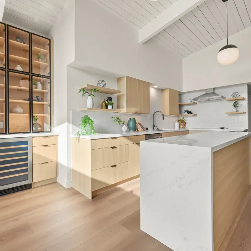A modern open-concept kitchen featuring light wood cabinets and a white marble waterfall-edge island. The space includes glass-front display cabinets with interior lighting, a built-in wine cooler, and floating shelves decorated with plants and dishware. The design showcases a vaulted white ceiling with exposed beams, pendant lighting, and a neutral color palette of white, light wood, and stainless steel accents.