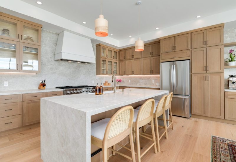 A modern kitchen featuring light wood cabinetry and a large white marble waterfall island with three woven cane-back bar stools. The space includes stainless steel appliances, a white range hood, pendant lighting, and white subway tile backsplash, all set against pale hardwood flooring that creates a warm, contemporary aesthetic.