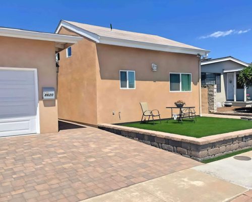 A single-story stucco home with beige/peach exterior walls and white trim, featuring a white garage door and multiple windows. The front yard showcases landscaping with artificial green turf, raised brick-bordered planting areas, and paver stone driveway under a clear blue sky. A patio set with table and chairs sits on the lawn area, with neighboring homes visible in the background.