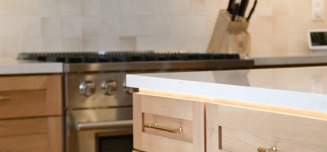 A modern kitchen interior featuring light wood cabinetry with brass pull handles and a white countertop. In the background, a stainless steel range sits beneath a dark countertop, with a wooden knife block visible on the counter. The kitchen has a clean, minimalist design with pale tile backsplash and warm natural wood tones throughout.