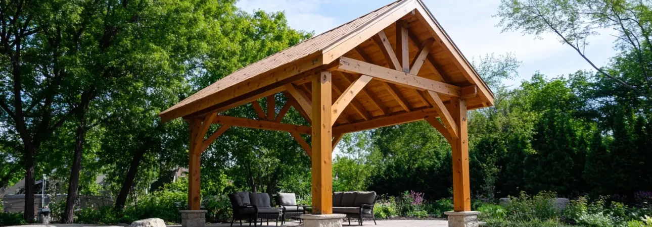 A timber frame pavilion with exposed wooden beams and a peaked roof stands in a landscaped backyard. The open-air structure shelters patio furniture on a concrete pad, surrounded by manicured lawn, decorative boulders, and lush green trees. The natural wood construction features traditional post-and-beam carpentry with diagonal bracing visible in the gable end.
