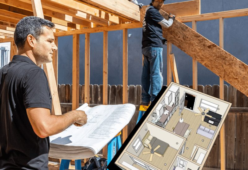 A construction supervisor in a black shirt reviews architectural blueprints on a work table at an active building site, where workers are installing wooden framing and plywood panels on a structure. A detailed floor plan model is visible on the table alongside the blueprints. The scene shows residential construction in progress with exposed wood framing against a dark exterior wall.