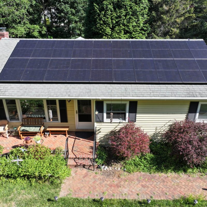An aerial view of a single-story ranch-style home with yellow siding and a large array of solar panels covering most of the roof. The house features a front porch with outdoor furniture and a brick patio area, surrounded by mature trees and landscaping including ornamental shrubs with reddish foliage. The solar panel installation appears professionally mounted on the shingled roof, demonstrating residential renewable energy implementation.