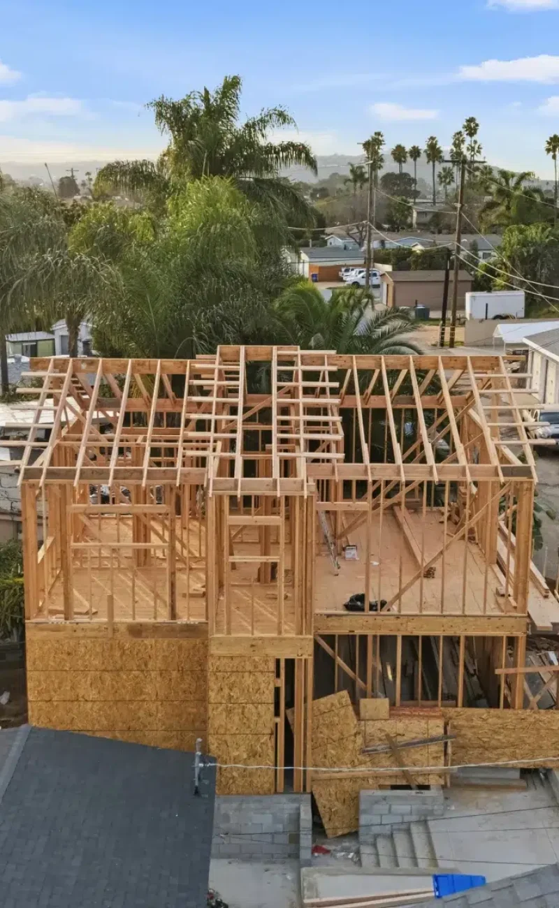 Aerial view of a two-story residential construction site showing the wooden frame and roof structure in progress. The house is surrounded by established neighborhood homes with palm trees and various vegetation visible throughout the suburban setting. Two vehicles are parked on the property, and the skeletal framework reveals the building's layout during the framing phase of construction.
