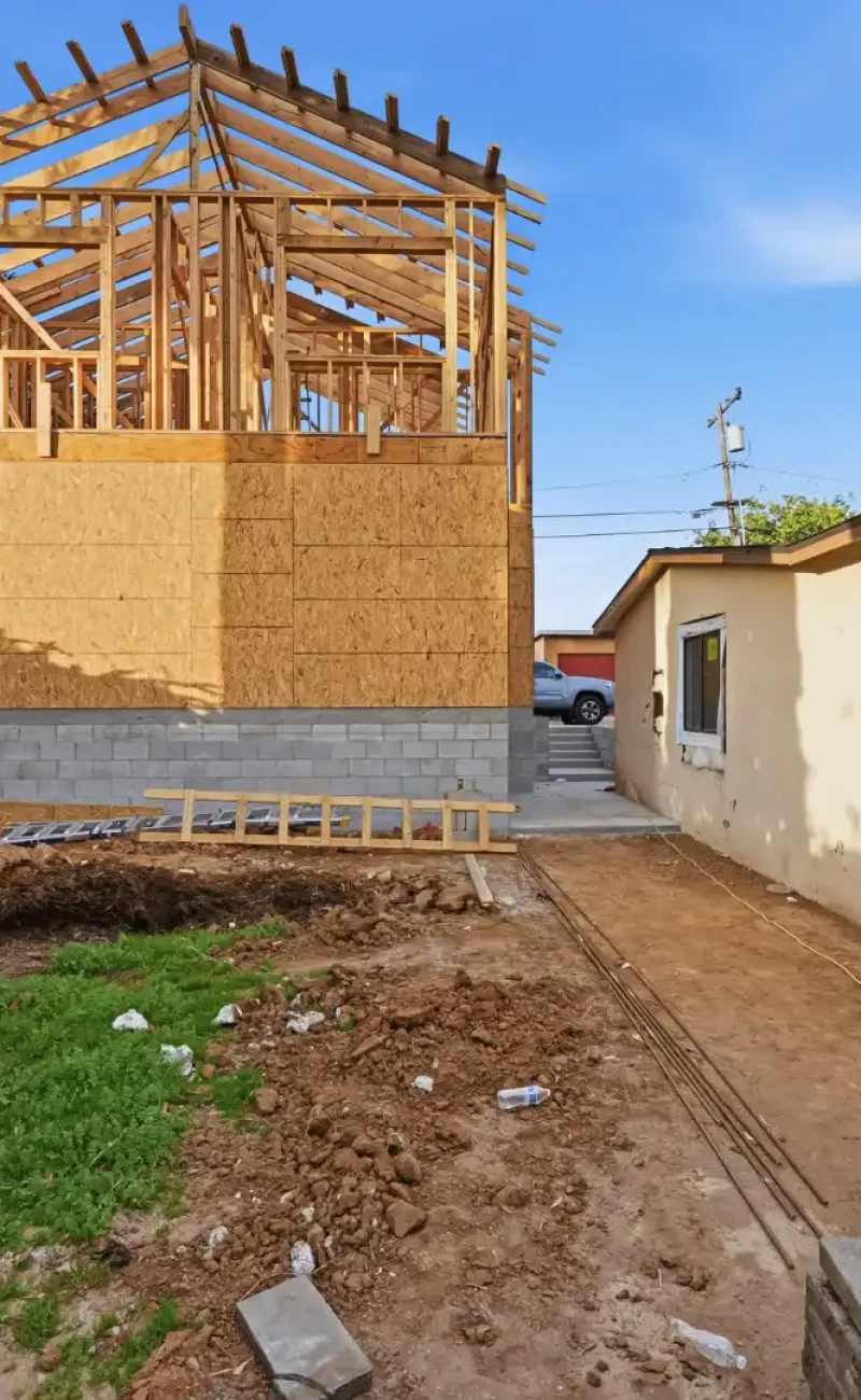 A two-story residential addition under construction in a backyard, showing wood frame structure with exposed roof joists sitting on a concrete block foundation. The new structure is positioned between palm trees on the left and an existing single-story beige house on the right, with disturbed dirt and patches of grass in the foreground indicating active construction work.