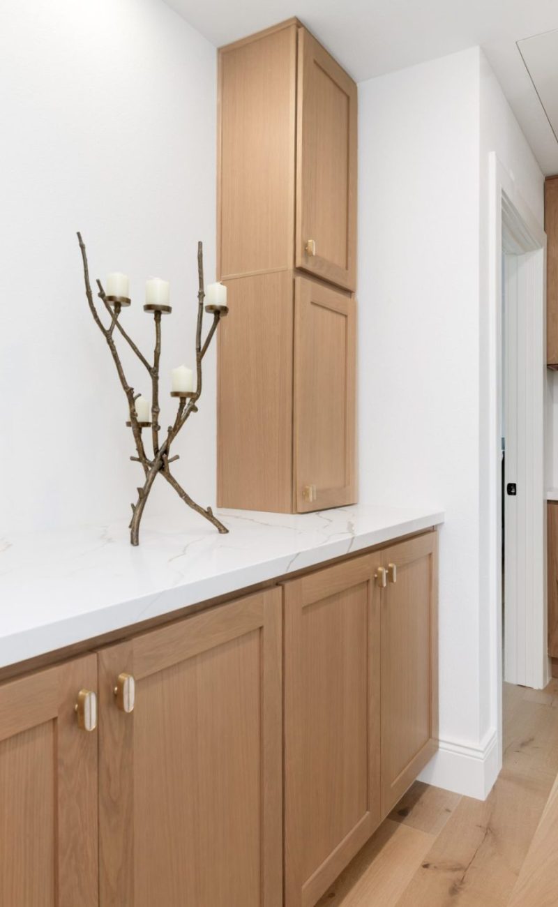 A modern hallway featuring light oak wood cabinetry with white marble countertops on both sides. The minimalist space has white walls, light hardwood flooring, and is decorated with a rustic branch candelabra and a vase of fresh flowers with red accents. Built-in storage cabinets extend to the ceiling, providing ample organization in this clean, contemporary interior.