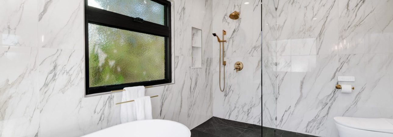 A modern luxury bathroom featuring floor-to-ceiling white marble walls with gray veining, a freestanding white soaking tub, and a frameless glass shower enclosure with gold fixtures. The space has dark slate-colored floor tiles creating dramatic contrast, recessed ceiling lights, and a black-framed frosted window providing natural light and privacy.