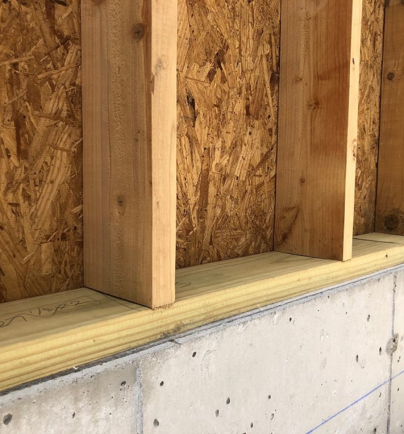 Interior view of a wood-framed structure under construction, showing exposed wall framing with OSB sheathing panels and dimensional lumber studs. The framing sits on a concrete foundation wall with visible anchor bolts. The raw construction materials and unfinished state indicate this is an active building project in the early framing stage.