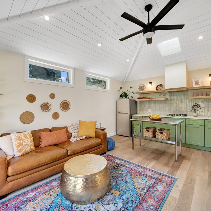 A bright modern ADU interior featuring vaulted shiplap ceilings with a black ceiling fan, combining a living area with a tan leather sofa and decorative wall baskets, and a kitchenette with sage green cabinets, white countertops, and stainless steel appliances. The space has light wood flooring, a colorful Persian-style rug, and large windows providing natural light. The open-concept design maximizes the small footprint with efficient storage and a cohesive bohemian-modern aesthetic.