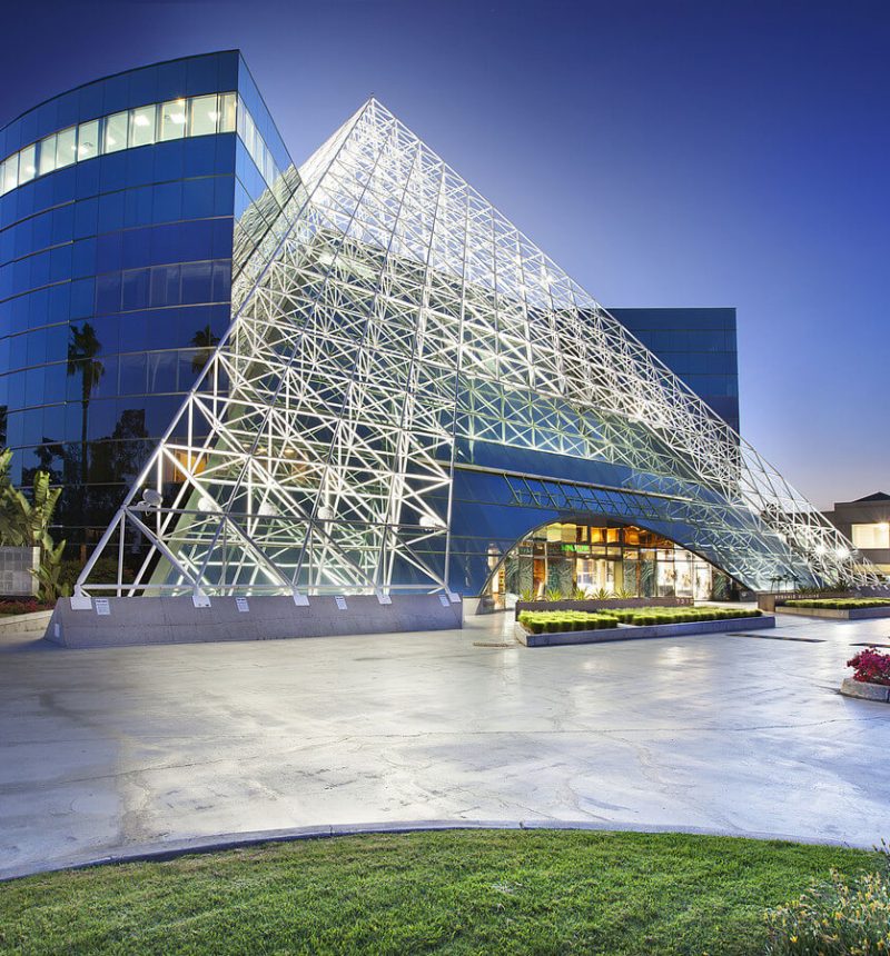 A modern commercial building complex photographed at dusk featuring a distinctive white geometric pyramid-shaped steel framework structure in front of curved and angular glass-clad office towers. The illuminated buildings and landscaping with palm trees, flowers, and manicured lawn create a dramatic twilight scene. The architectural composition showcases contemporary design with the contrasting structural elements of the skeletal pyramid against the sleek blue glass facades.