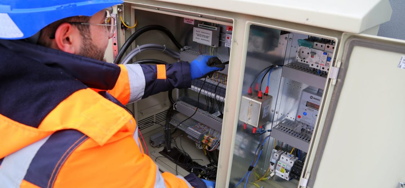 A technician wearing a blue hard hat and high-visibility orange and navy safety vest performs maintenance on an open electrical control panel. The worker, wearing blue gloves and safety glasses, reaches into the cabinet which contains circuit breakers, wiring, and various electrical components. The white industrial equipment cabinet has multiple compartments with organized electrical infrastructure visible inside.