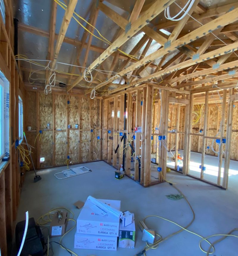 Interior view of a residential building under construction showing exposed wooden framing, ceiling joists, and electrical wiring throughout. The space features light-colored subflooring, OSB wall sheathing, and blue electrical junction boxes mounted on wall studs. Construction materials and boxes are visible on the floor, with natural light entering through window openings on the left side.