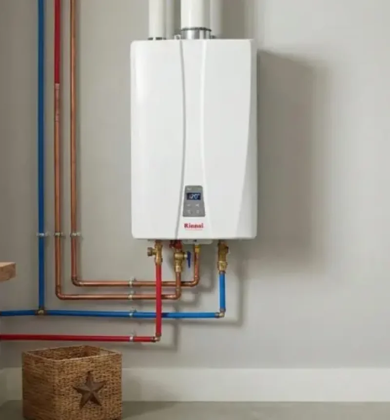 A white wall-mounted tankless water heater installed in a laundry or utility room, with red and blue PEX pipes connecting to the unit. The modern appliance features a digital display panel and is mounted on a light gray wall, with wooden shelving holding folded towels visible to the left and a wicker basket below.