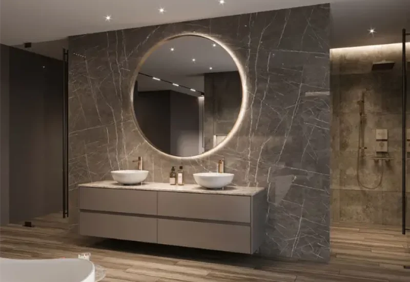 A luxurious modern bathroom featuring a double vanity with white vessel sinks mounted on a floating taupe cabinet. The focal point is a large circular illuminated mirror against a dramatic gray marble accent wall with white veining. The space includes wood-look tile flooring, recessed lighting, and a glass-enclosed shower visible in the background, creating an elegant spa-like atmosphere.