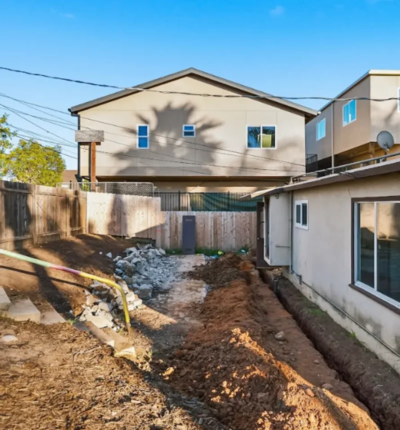 A residential construction site showing a two-story beige house with a gabled roof in the background, surrounded by neighboring homes. The foreground shows excavated dirt, exposed utility pipes including a yellow gas line, rocks, and construction debris scattered across the undeveloped yard. Wooden fencing separates the property from adjacent houses in this densely built neighborhood.
