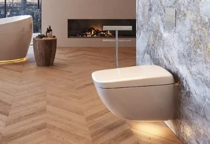 A modern wall-mounted white toilet with an integrated bidet function is mounted above light herringbone wood flooring in a luxurious bathroom. The toilet features ambient LED lighting underneath and is positioned near a marble accent wall and a contemporary linear fireplace. A wooden cylindrical side table completes the minimalist, spa-like aesthetic of the space.