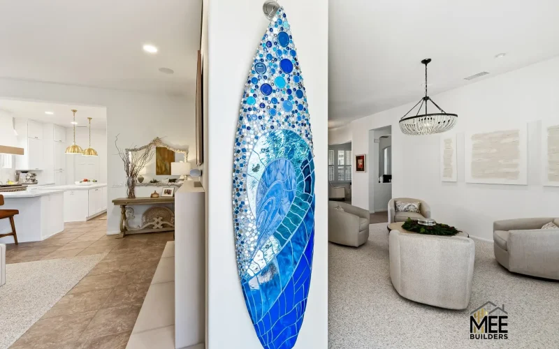 A decorative surfboard with blue mosaic tile artwork hangs vertically on a white wall between an open-plan kitchen and living room. The surfboard features various shades of blue tiles creating an ocean wave pattern, with circular bubble-like designs at the top transitioning to geometric patterns below. The modern coastal home interior showcases white cabinetry, beige furniture, and neutral tones throughout the space.