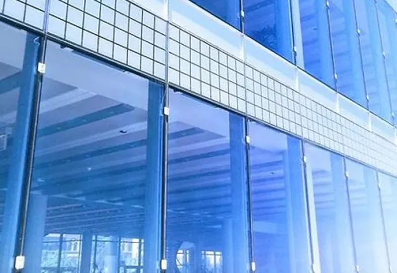 A modern commercial building facade featuring floor-to-ceiling glass curtain walls with blue-tinted windows and white metal framing. The architectural design showcases large transparent panels separated by vertical and horizontal metal mullions, with a distinctive horizontal band of smaller glass blocks or tiles running across the upper portion. The reflective glass surfaces mirror interior ceiling structures and adjacent buildings, creating a sleek contemporary aesthetic.