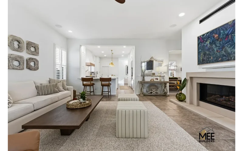 A modern open-concept living room featuring a beige sofa with striped pillows, dark wood coffee table, and two striped cube ottomans on plush carpeting. The space flows into a white kitchen with bar seating, while the living area showcases a fireplace with cream surround, botanical artwork above, and decorative wall accents. The room has a neutral color palette with white walls, recessed lighting, and natural textures throughout.