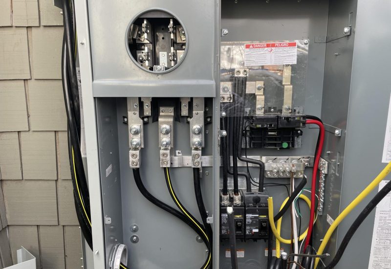 An open electrical meter base and service panel showing the main service entrance with three large conductors (two black, one yellow) connected to bus bars at the top. The right side contains circuit breakers and a danger warning label, with additional yellow and red conductors feeding into the panel. This is a typical residential or light commercial 200-amp electrical service installation mounted on an exterior wall with shingled siding.