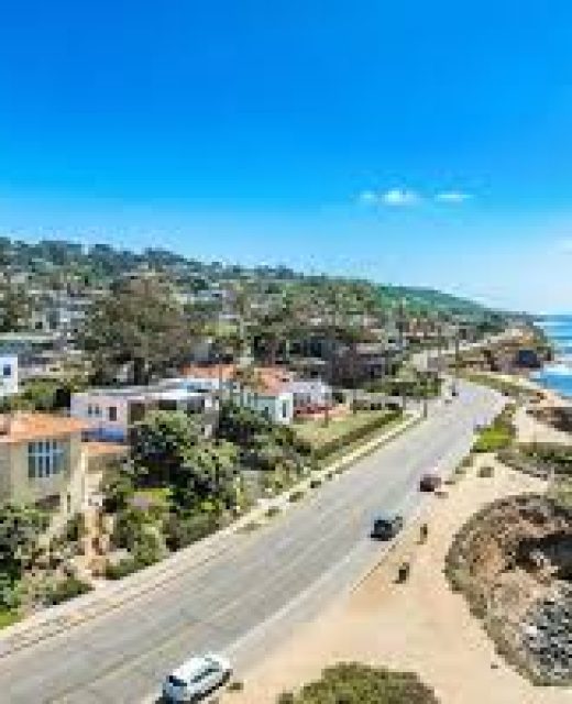 A scenic coastal view of a hillside community with residential homes along a curved oceanfront road. The image shows a mix of architecture nestled on terraced slopes, with the Pacific Ocean visible on the right side under a clear blue sky. The coastal highway runs parallel to the shoreline, with sandy areas and rocky outcroppings visible along the water's edge.