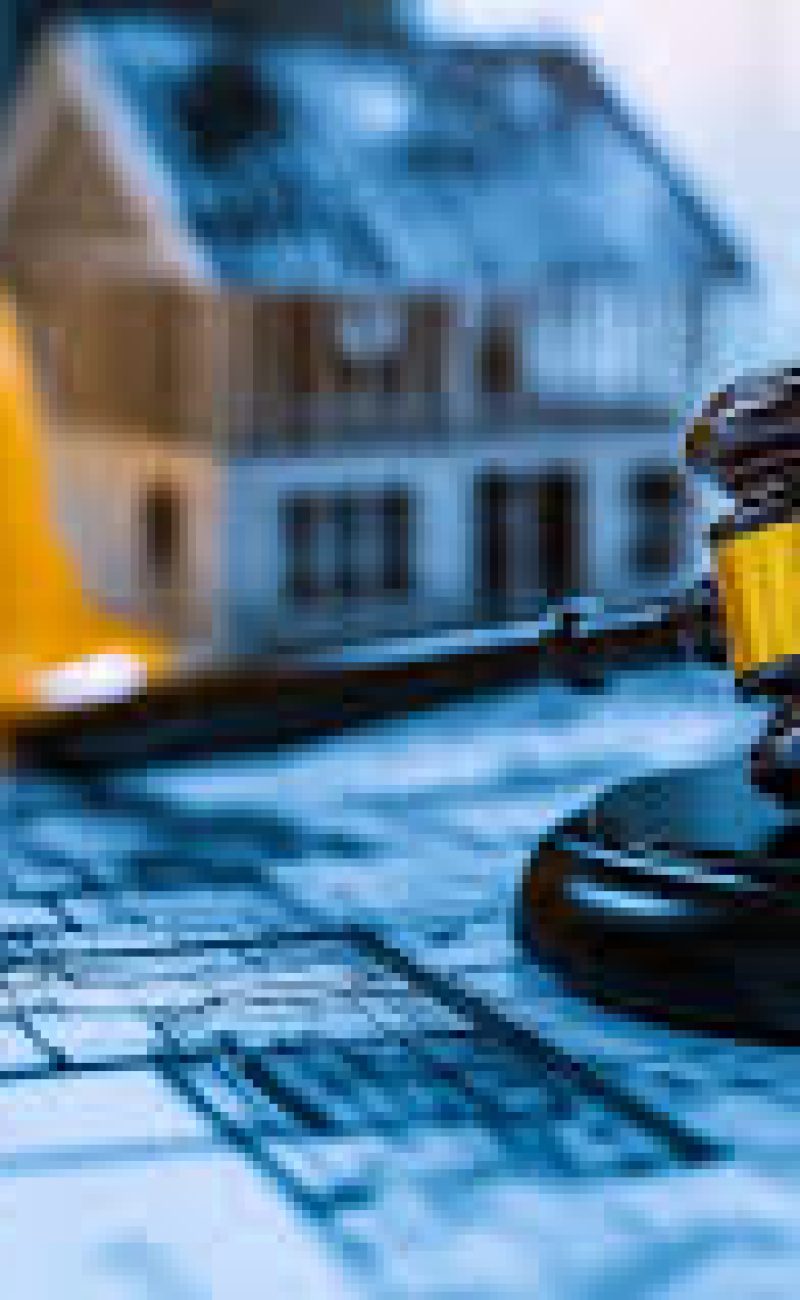 A judge's gavel rests on architectural blueprints alongside a yellow hard hat and a small house model, symbolizing construction law and real estate litigation. The composition suggests legal matters related to building projects, property disputes, or construction contract negotiations. The professional setting indicates the intersection of legal proceedings and the construction industry.