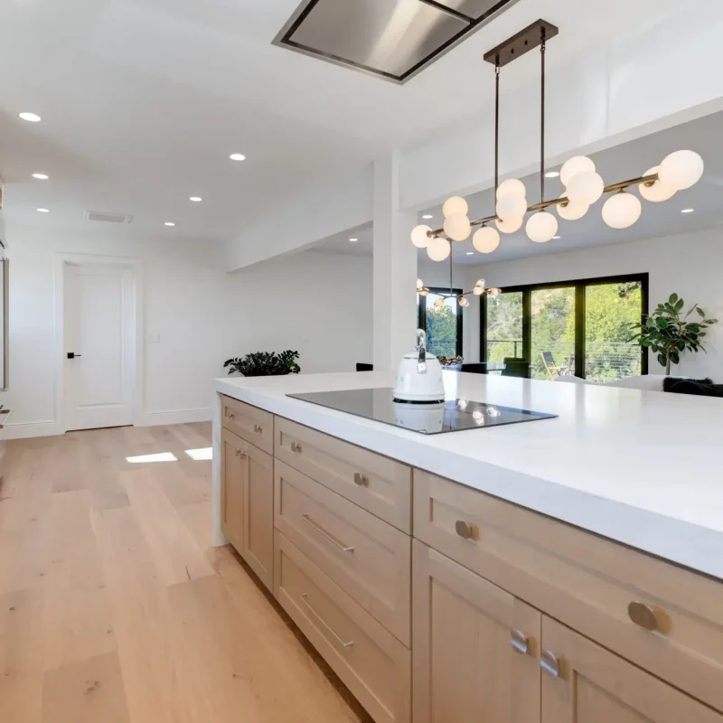 A modern open-concept kitchen featuring light wood cabinetry with white quartz countertops and a large center island with an induction cooktop. The space is illuminated by a contemporary chandelier with globe lights and recessed ceiling lights, while black-framed windows provide natural light and views of surrounding greenery. The kitchen showcases a minimalist design with light hardwood flooring and stainless steel appliances built into the left wall cabinetry.