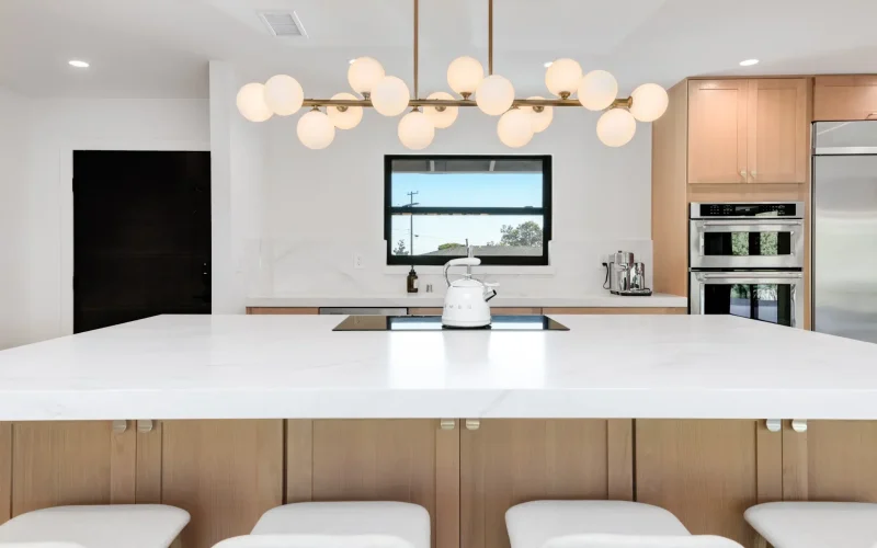 A modern kitchen featuring a large white island with light wood base cabinets and white upholstered bar stools. Above the island hangs a contemporary brass chandelier with multiple white globe lights. The kitchen includes light wood cabinetry, stainless steel appliances, a window backsplash, and a sleek black door, all set against white walls with a skylight visible above.