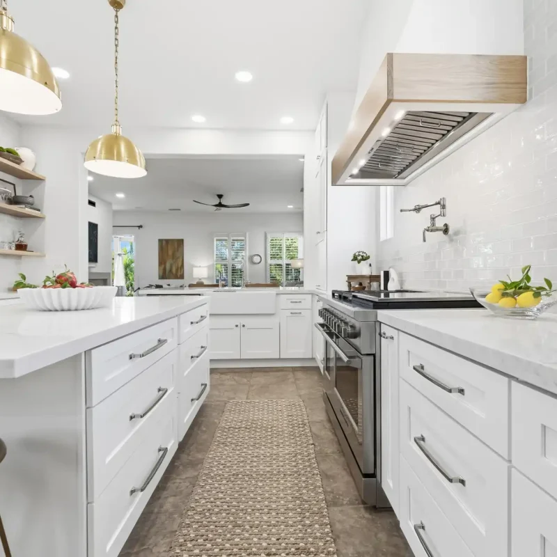 A modern white kitchen featuring white shaker-style cabinets, white quartz countertops, and stainless steel appliances. The space includes a kitchen island with seating, gold dome pendant lights, and a custom wood-wrapped range hood. The bright, open layout showcases clean lines, professional-grade appliances, and a neutral runner rug along the cooking zone.