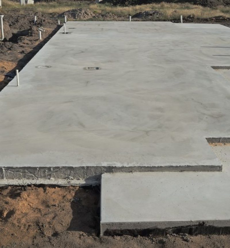 A freshly poured concrete foundation slab for a building under construction, with white PVC pipes protruding vertically from the surface at regular intervals for future plumbing connections. The slab features visible expansion joints and is surrounded by exposed earth and construction site debris. Wooden stakes and string lines are visible in the background, marking the perimeter of the construction area.