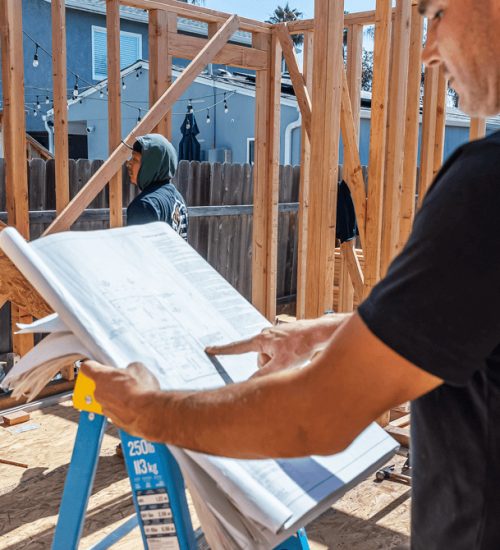 A construction worker in a black shirt reviews architectural blueprints at an active building site with wooden framing in progress. The blueprints are spread across a blue ladder while another worker in a hooded sweatshirt works on the frame structure in the background. A residential fence and neighboring buildings are visible behind the construction area.